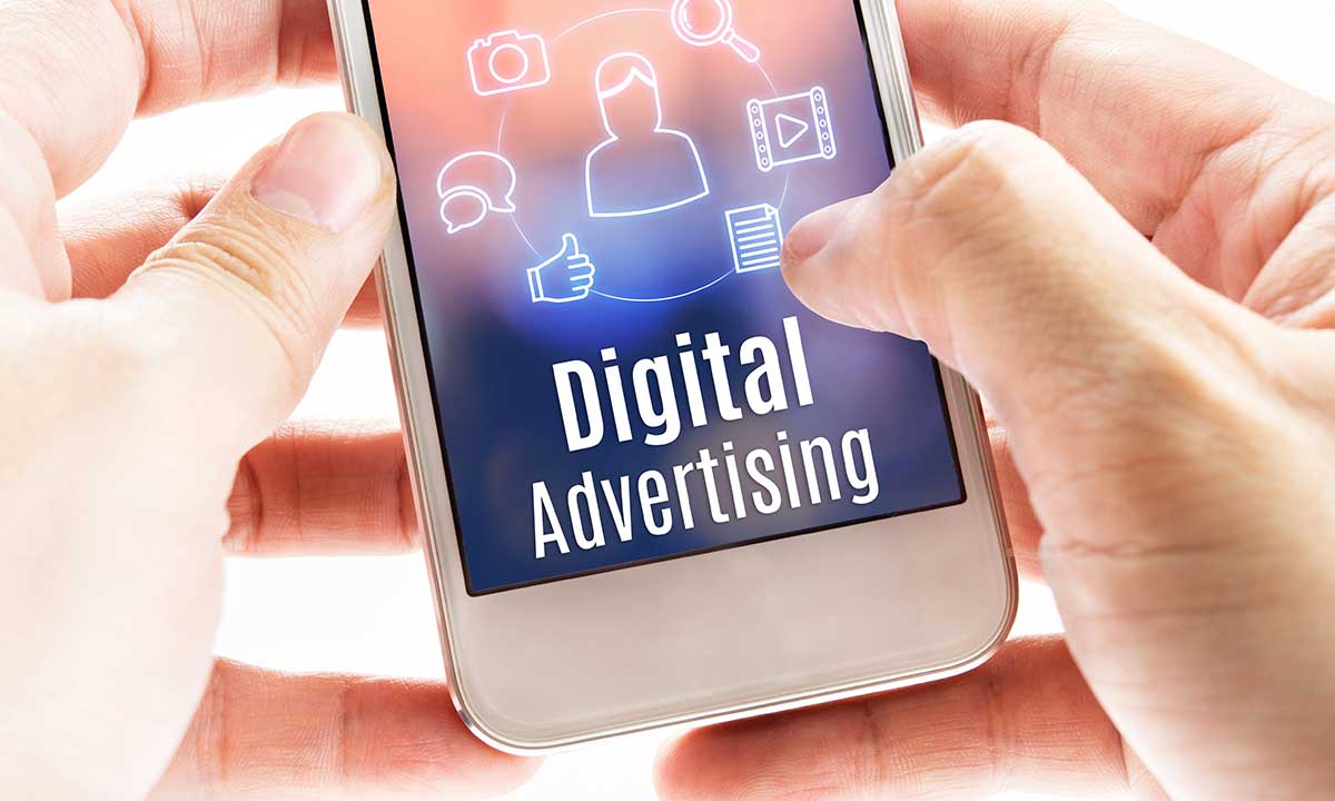 Hands holding a smartphone displaying 'Digital Advertising' text and icons related to online marketing such as thumbs up, camera, and messaging, illustrating the concept of digital advertising trends.