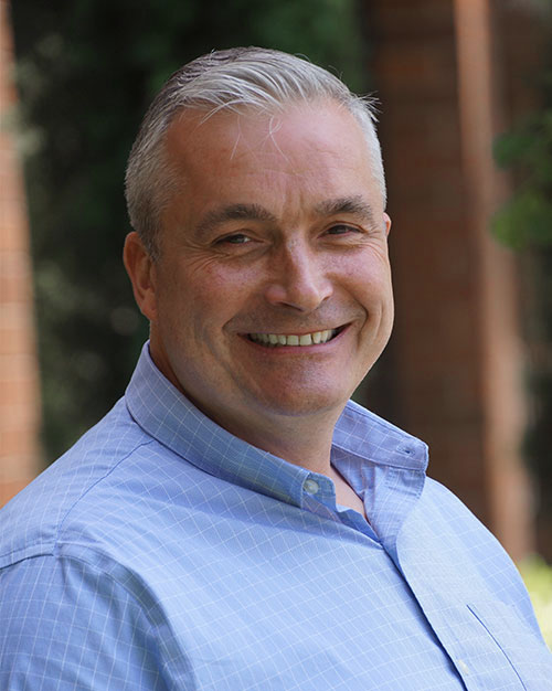 Portrait of Brendan Scott, a professional content creator, with a warm smile, dressed in a blue checked shirt against a natural outdoor background.