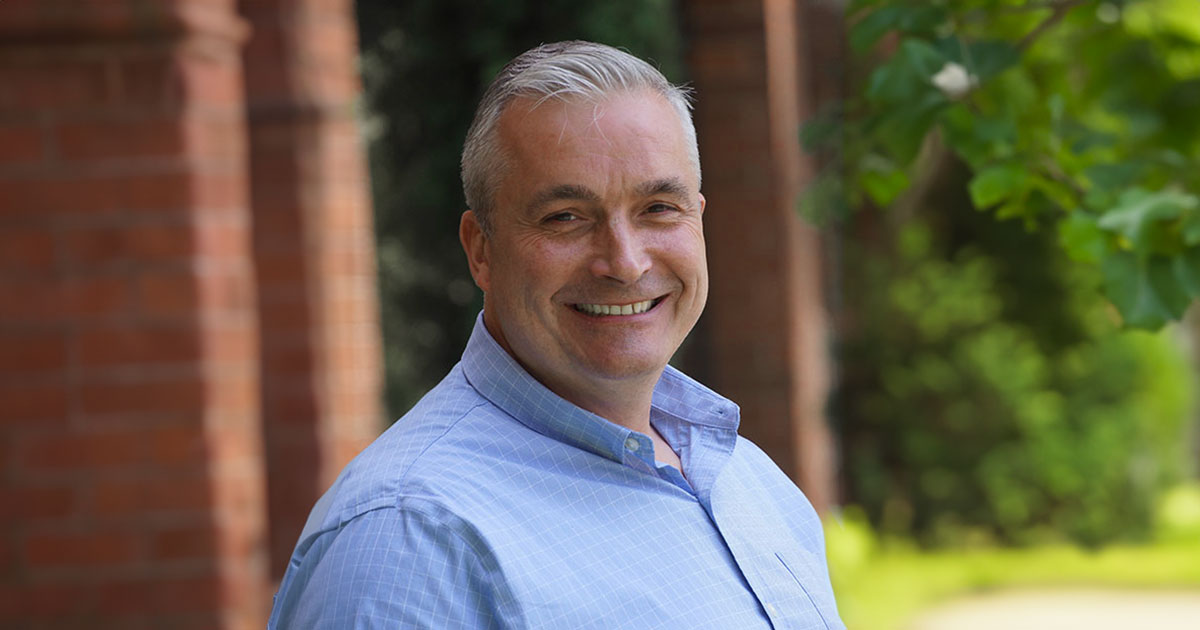 Brendan Scott posing confidently, wearing a blue checked shirt, with a serene brick building and greenery in the background, representing his grounded and approachable demeanor.